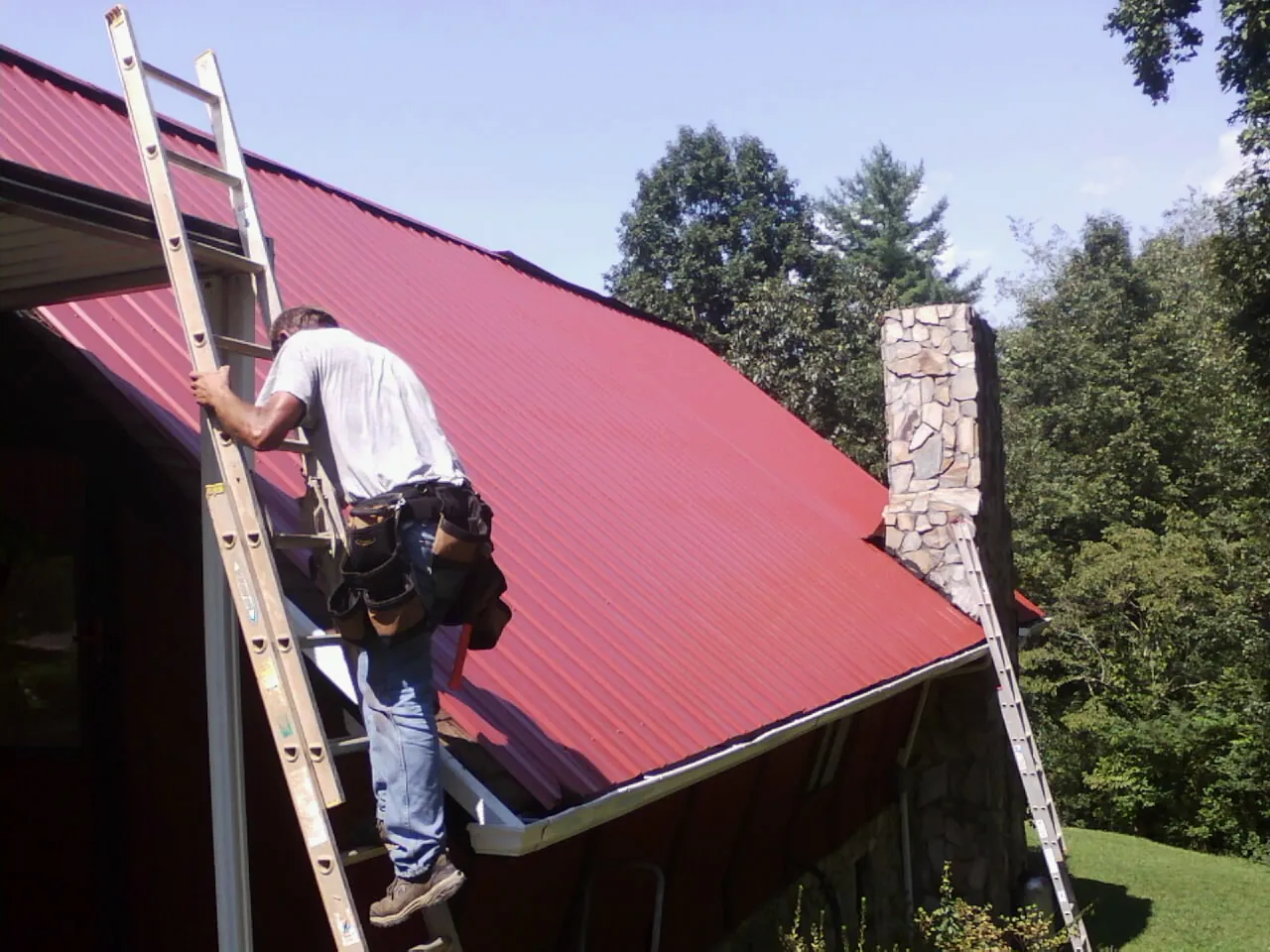 Roofer on a ladder beside a red metal roof and stone chimney — Soffit & Fascia Repair by Roofing Brothers in Rittman